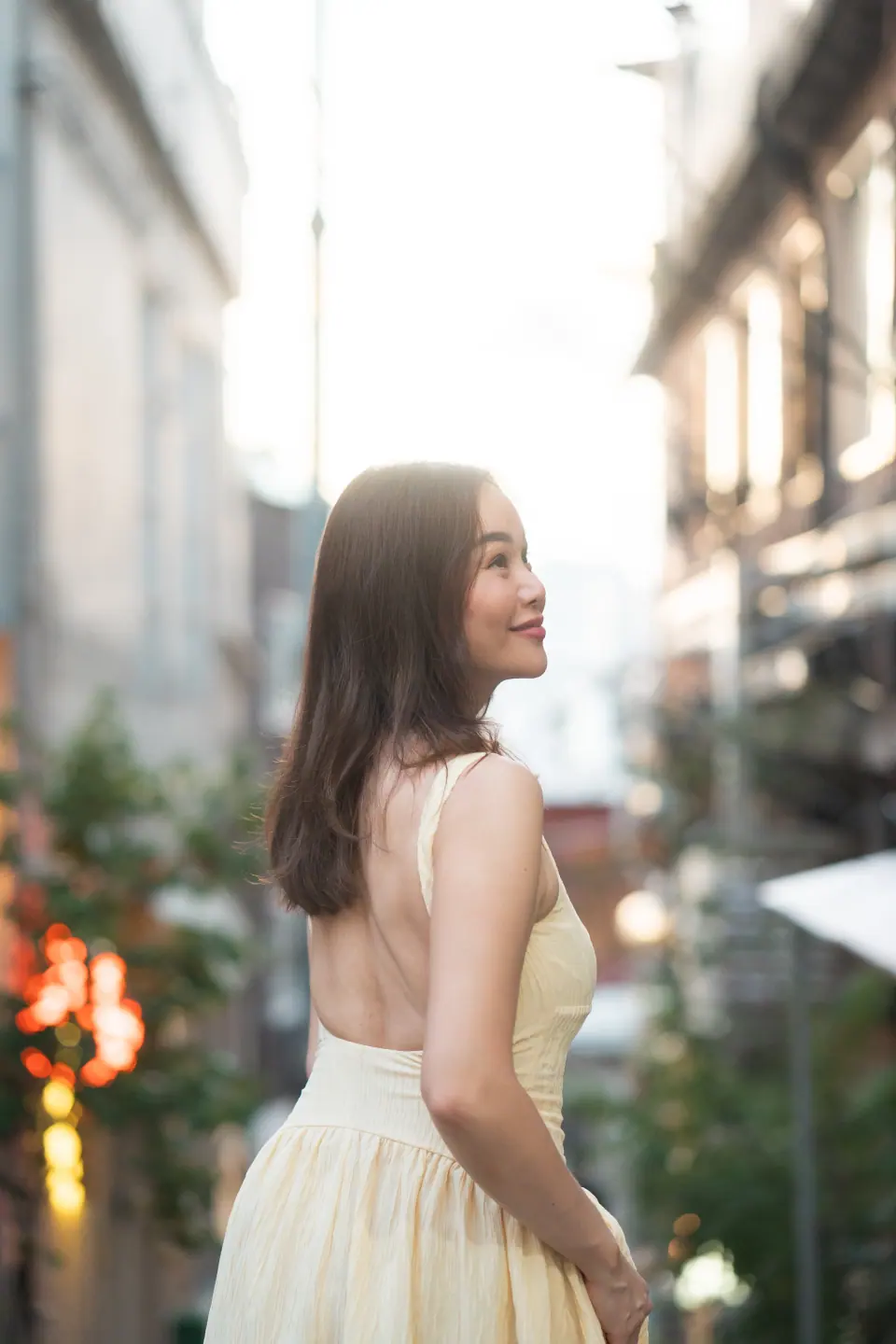 A woman in a cream dress smiling toward the light in a street