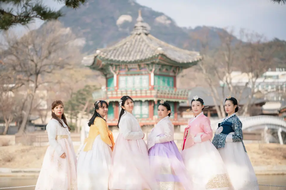 Women in hanbok in front of a palace pavilion, mountains behind